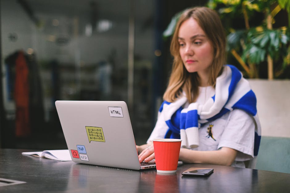 A young woman seated at an outdoor table engaged in gardening or landscaping work, with a silver laptop open in front of her displaying labels like HTML, CSS, and PHP stickers. The table holds a red p